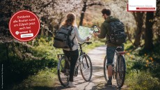 Zwei Menschen stehen mit ihren Fahrrädern auf einem Waldweg. Die linke Person hält eine Fahrradkarte in der Hand, die rechte ihr Handy.