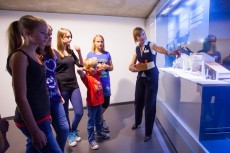 Eine Frau steht vor einer Vitrine und zeigt auf etwas. Vor ihr steht eine Gruppe Jugendlicher.