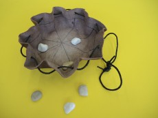 Auf einem gelben Untergrund liegt ein geöffnetes Säckchen mit einem eigenzeichneten Mühlebrett darin, daneben liegen Kieselsteine.