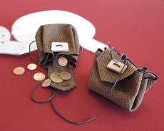 Auf einem roten Untergrund liegen zwei kleine Ledersäckchen. Das eine ist geöffnet und ein wenig Münzgeld fällt heraus.