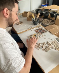 Ein junger Mann sitzt an einem Tisch und legt aus kleinen Steinchen ein Mosaik.